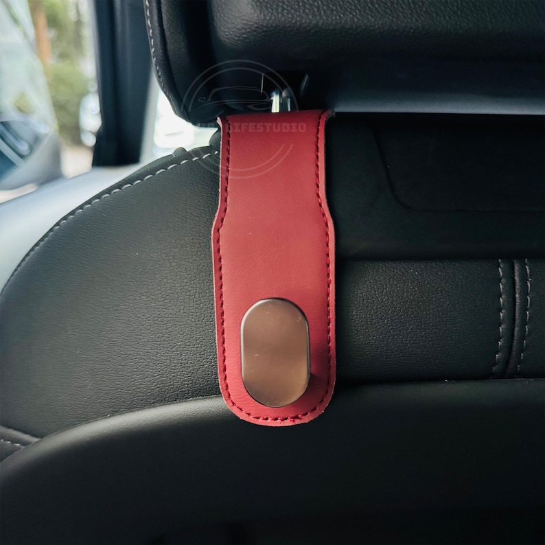 May include: A red leather car seat headrest hook with a metallic oval hook. The hook is attached to a black car seat. The stitching on the hook is red.