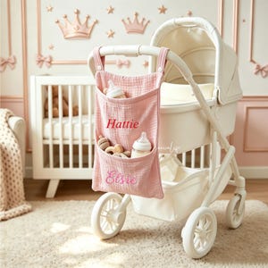 May include: A pink muslin fabric stroller organizer hanging from a white baby carriage. The organizer features two pockets with the embroidered names Hattie and Elsie in red and pink thread, containing two baby bottles and small wooden toys.