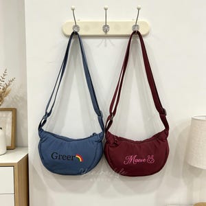 May include: Two crescent-shaped crossbody bags with adjustable straps, one in slate blue featuring the name Greer and a rainbow icon, and one in deep burgundy featuring the name Maeve and a small rabbit icon, both with top zipper closures.