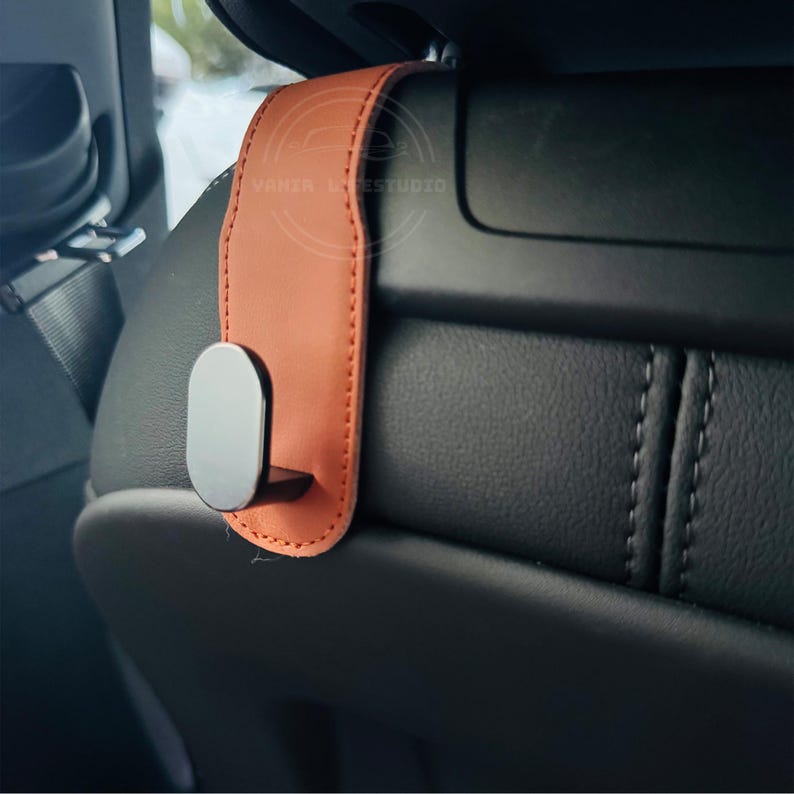 May include: A car seat headrest hook with a brown leather strap and a silver-colored hook. The hook is designed to hang items in a vehicle. The leather strap is stitched with a contrasting thread.