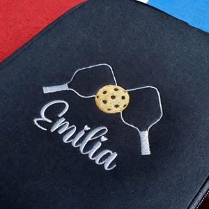 Op de afbeelding: Een zwarte pickleball paddle case met de naam "Emilia" geborduurd in witte letters. Het ontwerp omvat witte pickleball paddles en een gele pickleball. De case sluit met een rits.