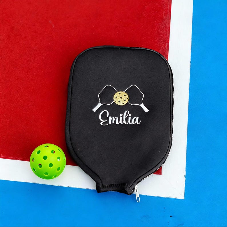 Op de afbeelding: Zwarte pickleball paddle cover met een rits, wit geborduurd pickleball paddle en bal ontwerp, en de naam "Emilia" in witte letters. Een groene pickleball met gaten ligt naast de cover op een rood en blauw oppervlak.