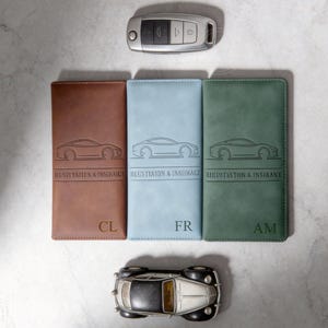 May include: Three leather car registration and insurance holders in brown, light blue, and green, each with a car silhouette and the text "REGISTRATION & INSURANCE". The holders have initials "CL", "FR", and "AM". A car key and a toy car are also present.