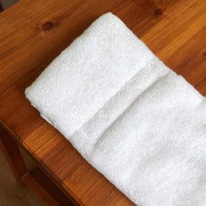 May include: A folded white bath towel with a textured terry cloth surface. The towel is rectangular in shape and features a clean, solid white color throughout its entire fabric construction, showing a soft and absorbent material texture.
