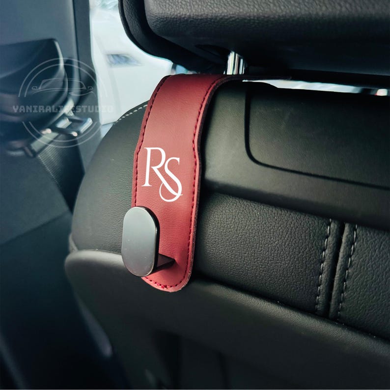May include: A burgundy car seat headrest hook with the white initials "RS" printed on it. The hook is made of a leather-like material and has a black metal hook. The hook is attached to a black car seat.