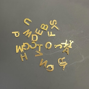 Peut inclure: Un ensemble de breloques en m&eacute;tal dor&eacute; en forme de lettres, &eacute;parpill&eacute;es sur une surface grise. Les lettres comprennent A, B, C, D, E, F, G, H, I, J, K, L, M, N, O, P, Q, R, S, T, U, V, W, X, Y et Z.