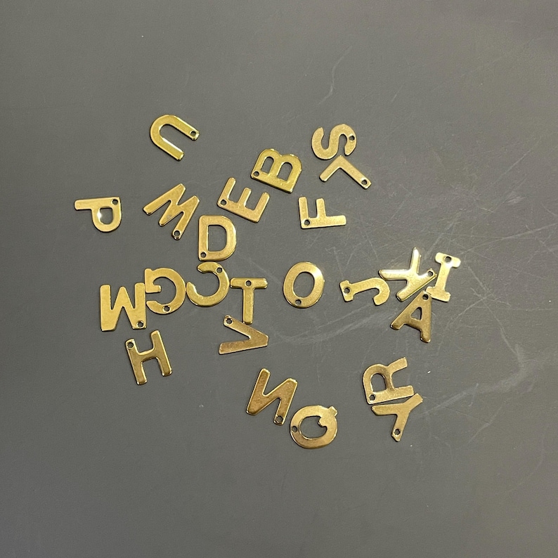 Peut inclure: Un ensemble de breloques en m&eacute;tal dor&eacute; en forme de lettres, &eacute;parpill&eacute;es sur une surface grise. Les lettres comprennent A, B, C, D, E, F, G, H, I, J, K, L, M, N, O, P, Q, R, S, T, U, V, W, X, Y et Z.
