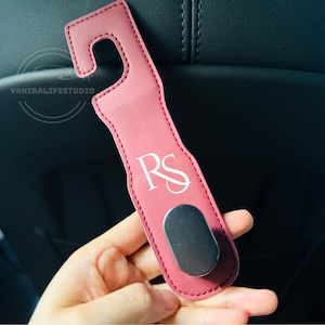 May include: A pink car seat hook with a curved top and a silver magnetic base. The hook has the initials "RS" in white script. The hook is made of a leather-like material.