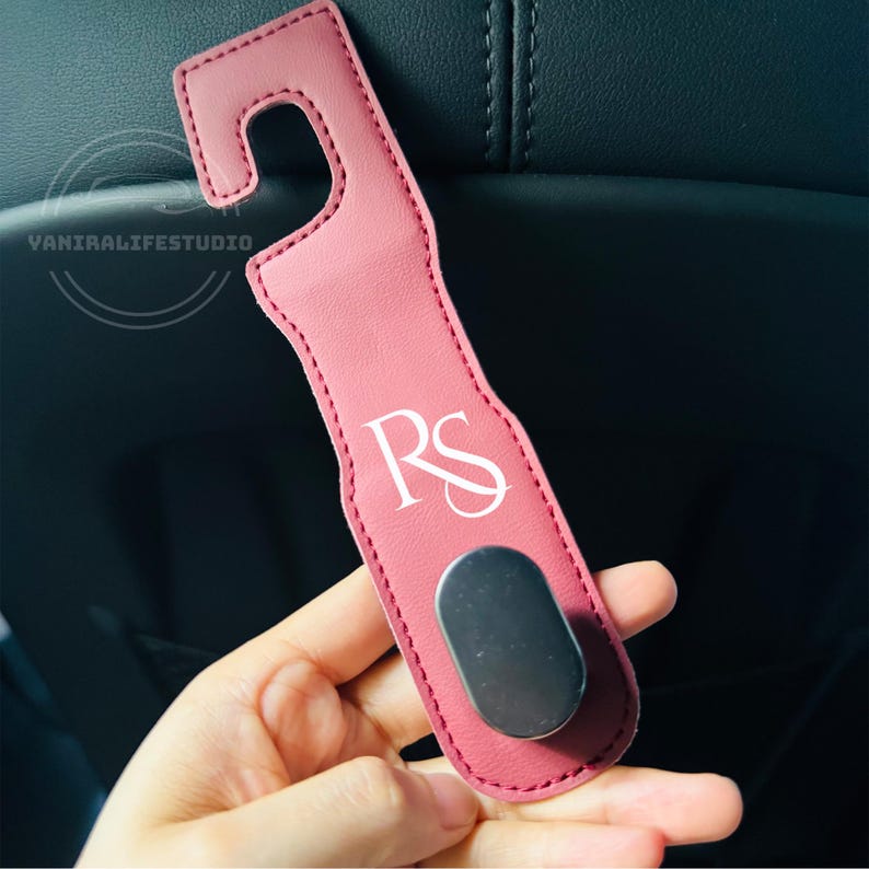 May include: A pink car seat hook with a curved top and a silver magnetic base. The hook has the initials "RS" in white script. The hook is made of a leather-like material.