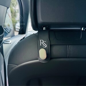 May include: Black leather car seat hook with a silver-colored hook and the initials "RS" in white. The hook is designed to hang from the headrest of a car seat.