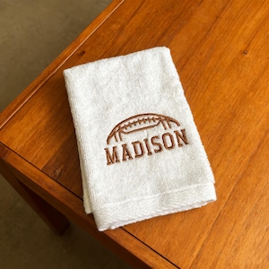 May include: A white rectangular hand towel featuring brown embroidery. The design includes a graphic of an American football positioned above the name MADISON, which is stitched in a bold, capitalized, brown sans-serif font.