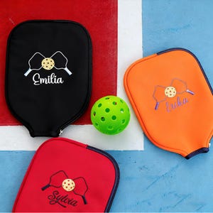May include: Three pickleball paddle covers in black, red, and orange, each with a personalized name embroidered on them. A green pickleball sits in the center. The covers feature a design of two paddles and a ball.