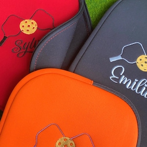 Op de afbeelding: Close-up van drie pickleball paddle covers in rood, oranje en zwart. Elke cover heeft een geborduurd pickleball- en paddle-ontwerp, met namen geborduurd in cursief. De covers hebben zwarte ritsen en staan op een groene achtergrond.