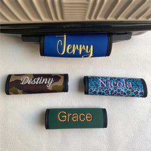 May include: A selection of luggage handle wraps. The wraps feature embroidered names: "Jerry" on blue, "Destiny" on camouflage, "Nicola" on a teal leopard print, and "Grace" on green. Each wrap has a black border.