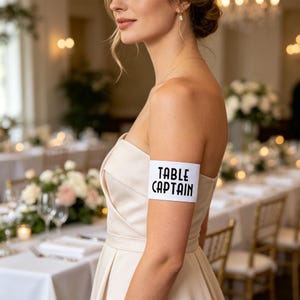以下が含まれることがあります： 太字のサンセリフ体で「TABLE CAPTAIN」と黒い文字がプリントされた白いアームバンド。上腕部に装着するタイプで、滑らかで不透明な素材を使用しており、端が真っ直ぐな長方形のデザインが特徴です。