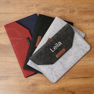Könnte beinhalten: Eine Sammlung von Laptop-Hüllen aus Filz in verschiedenen Farben: Rot, Marineblau, Schwarz und Grau. Jede Hülle hat einen Lederakzent und einen Klappenverschluss. Eine Hülle ist mit dem Namen "Leila" personalisiert.