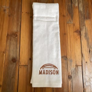 May include: A white hand towel featuring an embroidered brown football graphic positioned above the name MADISON in brown capital letters. The towel has a folded top edge and a soft, textured fabric surface.