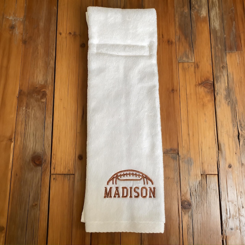 May include: A white hand towel featuring an embroidered brown football graphic positioned above the name MADISON in brown capital letters. The towel has a folded top edge and a soft, textured fabric surface.