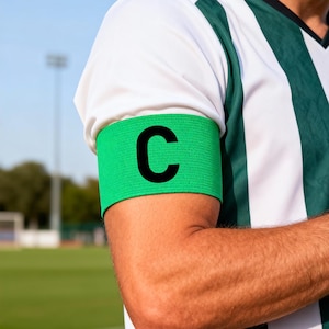 Pode incluir: Uma braçadeira de capitão verde com um grande "C" preto no braço de uma pessoa. A braçadeira é usada sobre uma camisa listrada branca e verde. O fundo mostra um campo verde e um céu limpo.