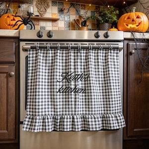 May include: A black and white gingham check oven curtain featuring a ruffled bottom hem and the embroidered text Keith's kitchen in black script. The curtain hangs from a metal rod attached to the front of a stainless steel oven door.