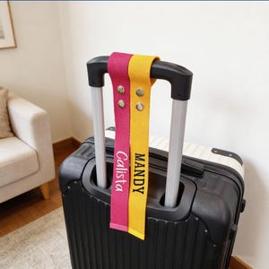 May include: A black suitcase with a retractable handle features two personalized luggage tags. One tag is fuchsia with the name "Calista," and the other is yellow with "MANDY." Both tags have metal snap closures.