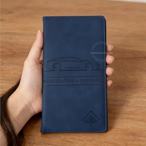 May include: A navy blue registration and insurance holder with a car outline and the text "REGISTRATION & INSURANCE". The holder has a logo at the bottom and is held in a hand.