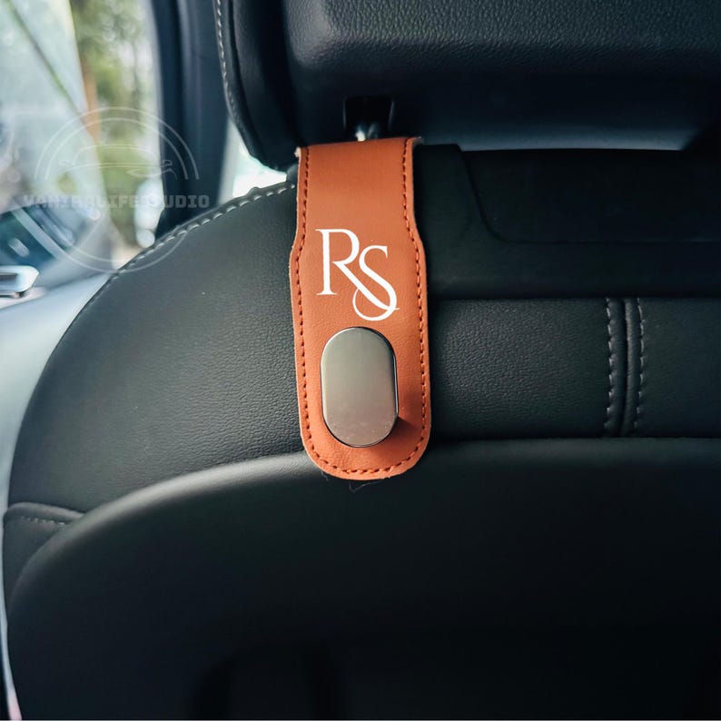 May include: A brown leather car seat headrest hook with the white initials "RS" printed on it. The hook has a silver-colored metal clip at the bottom. The hook is attached to a black car seat.