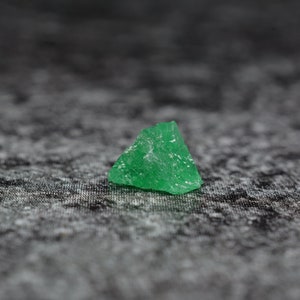 Könnte beinhalten: Ein einzelner, dreieckiger, grüner Edelstein mit einer rauen, facettierten Oberfläche. Der Edelstein liegt auf einer dunkelgrauen, strukturierten Oberfläche.