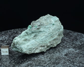 May include: A rough, gray-green rock with a few small, bright green emeralds embedded in it. The rock is sitting on a dark gray surface with a 1 cm ruler for scale.