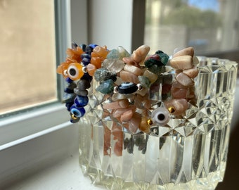 May include: A glass jar filled with a variety of colourful gemstone beads and charms, including blue, green, orange, and brown stones, and blue and white evil eye charms.