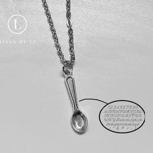 May include: A silver-toned spoon pendant hangs from a delicate chain. The image includes the text "RINGS BY LU" with a circular logo and an oval chart displaying uppercase and lowercase letters, numbers, and symbols for engraving reference.