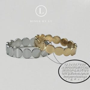 Op de afbeelding: Twee ringen met cirkelvormige schakels, één zilverkleurig en één goudkleurig, met een herhalend schijfpatroon. Een ovale afbeelding toont een lettertypekaart met cijfers 0-9, hoofdletters en kleine letters, en symbolen. Tekst RINGS BY LU.