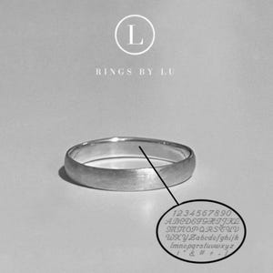 Könnte beinhalten: Ein silberfarbener Bandring mit gebürstetem, mattem Finish. Über dem Ring steht der Text L RINGS BY LU. Eine Linie führt vom Ring zu einer ovalen Grafik, die eine Tabelle der verfügbaren Zeichen zeigt, einschließlich der Zahlen 0-9, Groß- und Kleinbuchstaben sowie Symbole wie & # + - .