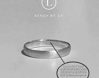 Puede incluir: Un anillo de banda plateado con un acabado mate cepillado. Sobre el anillo, el texto dice L RINGS BY LU. Una línea señala desde el anillo hacia un gráfico ovalado que muestra una tabla de caracteres disponibles, incluyendo números 0-9, letras mayúsculas y minúsculas, y símbolos como & # + - .