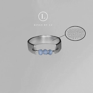 May include: A silver ring with a thin band and three light blue beads attached to a metal bar. The ring is from the brand "Rings by Lu".