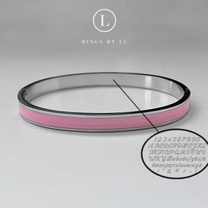 Puede incluir: Una pulsera de brazalete plateada con una banda central de esmalte rosa. La pulsera tiene un acabado pulido y un cierre de bisagra. Un primer plano muestra letras y números grabados. El texto "RINGS BY LU" está en la parte superior.