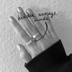 Könnte beinhalten: Zwei silberne Ringe werden an einer Hand mit manikürten Nägeln getragen. Die Ringe sind einfache, flache Bänder. Der Text "hidden message inside" steht über den Ringen mit einem Pfeil, der auf sie zeigt. Die Person trägt einen schwarzen Pullover.