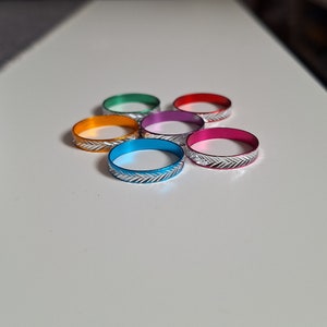 Könnte beinhalten: Sechs farbige Metallringe in Orange, Grün, Rot, Pink, Lila und Blau. Jeder Ring hat ein silbernes Zickzackmuster am Rand. Die Ringe sind kreisförmig auf einer weißen Oberfläche angeordnet.