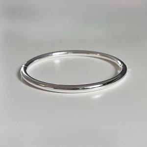 May include: A polished silver bangle bracelet with a smooth, rounded surface and a circular shape. The metal has a reflective finish that catches the light, creating bright highlights along the curve of the band. The bracelet is a simple, solid loop.