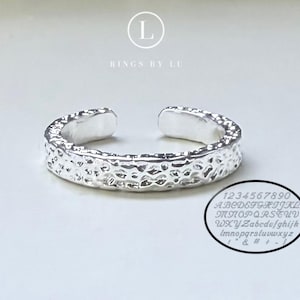 Könnte beinhalten: Ein silberfarbener offener Ring mit einer texturierten, gehämmerten Oberfläche. Über dem Ring steht der Text L RINGS BY LU. Unten rechts zeigt eine ovale Grafik die Zahlen 1234567890, das Alphabet in Groß- und Kleinbuchstaben sowie verschiedene Symbole.