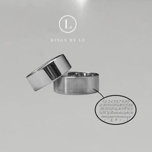 Op de afbeelding: Twee zilveren ringen, de ene iets gekanteld bovenop de andere, tegen een lichtgrijze achtergrond. De ringen zijn breedbandig en hebben een gepolijst, reflecterend oppervlak. Een kleine, cirkelvormige afbeelding toont cijfers, letters en symbolen.