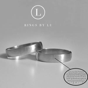 Puede incluir: Dos anillos de plata con un acabado de metal cepillado se muestran sobre una superficie gris claro. Los anillos están uno al lado del otro, con un primer plano que muestra una muestra de letras y números grabados. El logotipo de la marca "Rings by Lu" está en la parte superior.
