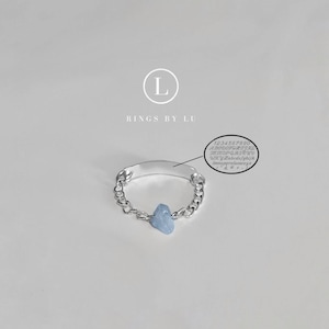 Puede incluir: Un anillo de cadena de plata con un dije de piedra azul claro. El anillo tiene una etiqueta de plata en blanco con el texto "Rings by Lu" arriba.