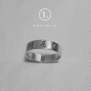 Könnte beinhalten: Ein silberner Ring mit der Inschrift "H & L" auf der Band. Der Ring ist in einem einfachen, minimalistischen Design gehalten.