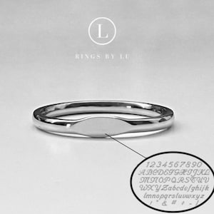 May include: A polished silver ring with an oval center. The ring is displayed on a light gray background. A small inset shows a sample of the available engraved fonts, including numbers, letters, and symbols. The text "Rings by Lu" is above the ring.