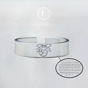Peut inclure: Une bague ouverte de couleur argentée avec un motif d'abeille gravé au centre. Au-dessus de la bague, le texte indique L RINGS BY LU. Sous la bague, un graphique ovale affiche un alphabet complet en majuscules et minuscules, les chiffres 0-9 et des symboles.