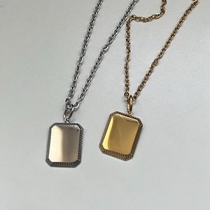 Può includere: Due collane con pendenti rettangolari. Una è color argento con una catena argentata, l'altra è color oro con una catena dorata. Entrambi i pendenti hanno un bordo strutturato e un centro liscio e riflettente.