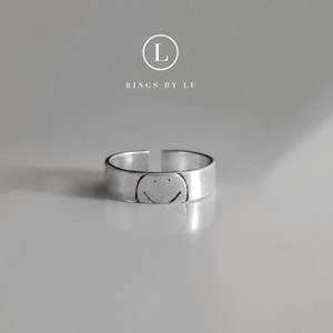 Könnte beinhalten: Ein silberfarbener offener Ring mit einem eingravierten Smiley-Gesicht. Über dem Ring befindet sich der Text L RINGS BY LU in einem weißen kreisförmigen Logo. Der Ring hat eine glatte, metallische Oberfläche und eine sichtbare Öffnung an der Rückseite.