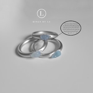 Könnte beinhalten: Drei silberne Ringe mit hellblauen Steinen, die in die Bänder eingefasst sind. Die Ringe sind übereinander gestapelt. Der Text "Rings by Lu" ist über den Ringen sichtbar.
