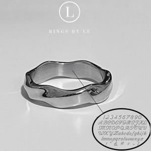 Può includere: Un anello argentato dal design ondulato. L'anello ha una superficie liscia e lucida e una forma unica e organica. L'immagine include anche un primo piano di lettere e numeri incisi.